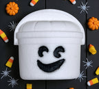 White foam bucket with ghost face design on a Halloween-themed background with pumpkins and candy corn.